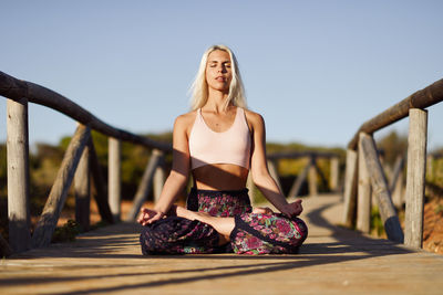 Full length of young woman meditating while sitting on boardwalk against sky