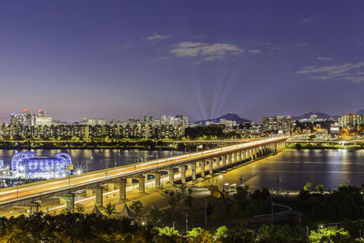 Illuminated bridge over river by buildings against sky at night