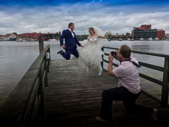 Male photographer photographing wedding couple jumping at pier over lake