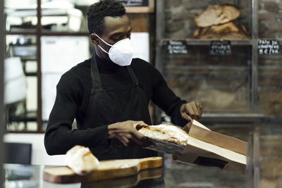 Male owner wearing face mask packing bread in paper bag while working at bakery