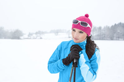 Thoughtful woman holding ski poles while standing on snow covered field