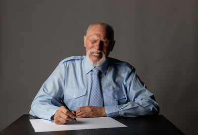 Senior businessman writing in paper and making face while sitting on table against gray background