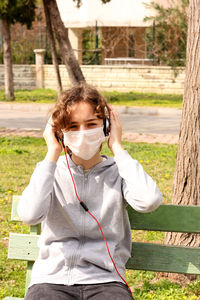 Young teenager girl with a medical mask is listening to music with headphones in park. 