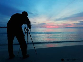 Rear view of man photographing while standing on shore at beach during sunset