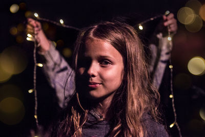 Close-up portrait of smiling girl against illuminated string lights at night