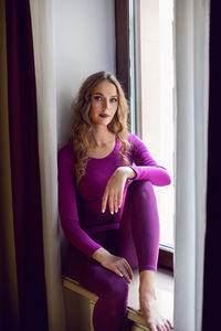 Young tall woman is sitting on windowsill in a hotel near the window in tight purple yoga clothes