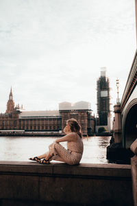 Woman sitting on bridge over river in city against sky