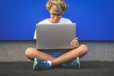 Boy using laptop while sitting against wall