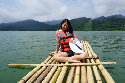 Smiling young woman sitting on wooden raft amidst lake against sky