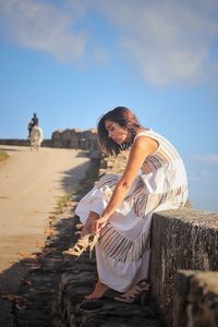 Side view of woman wearing high heels while sitting on retaining wall against sky