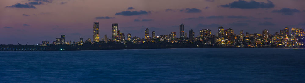 Illuminated city by sea against sky during sunset