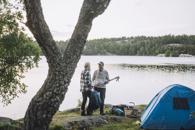 Mature man talking to friend standing by lake during camping