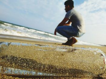 Close-up of bottle with man in background crouching at beach