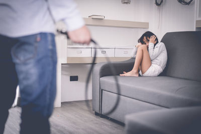 Midsection of young man holding belt while scared daughter sitting on sofa at home