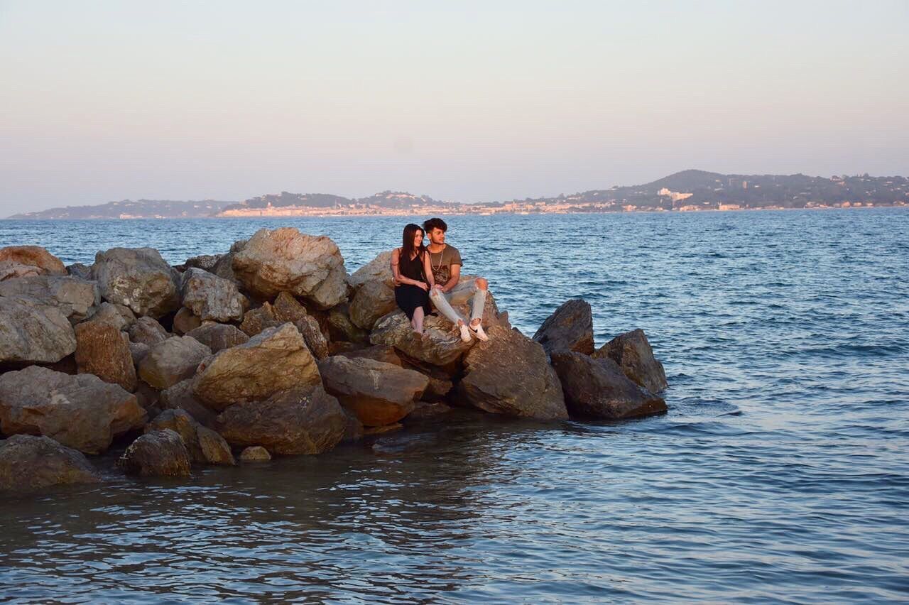 YOUNG WOMAN SITTING ON ROCK IN SEA AGAINST SKY