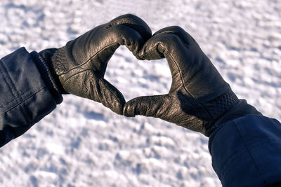 Male hands in black gloves are folded in shape of heart against background of white snow