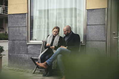 Full length of mid adult business people using smart phone on bench against building