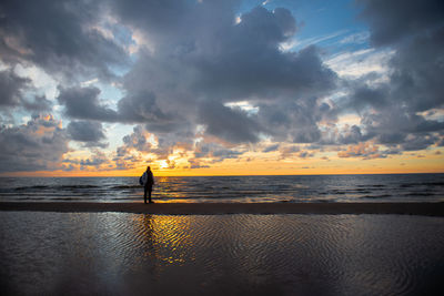 Man standing at beach against cloudy sky during sunset