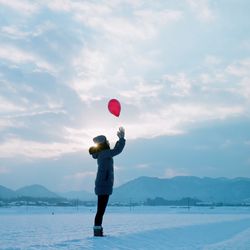 Side view of girl in warm clothing catching red balloon while standing on snowy field