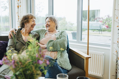 Smiling elderly women sitting with arm around while looking at each other on sofa against window at retirement home