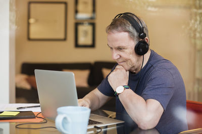 Senior man listening music while using laptop seen through glass window