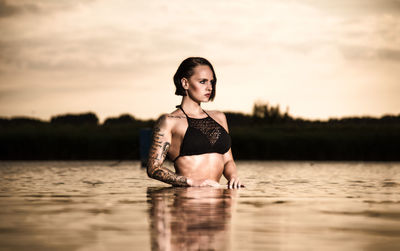 Young woman looking away standing in lake against sky during sunset