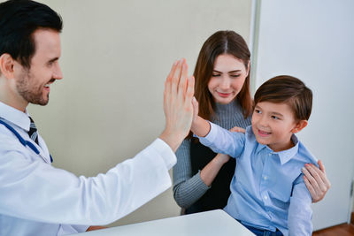 Cute boy sitting on lap of mother while giving high-five to doctor in hospital