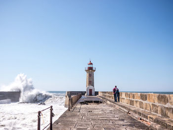 Lighthouse on sea by building against clear blue sky