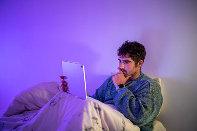 Man using digital tablet sitting on bed against wall at home