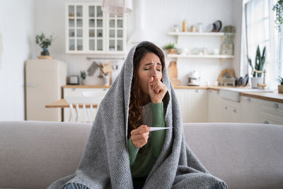 Sick woman coughing, measuring temperature with thermometer, sitting on sofa wrapped in blanket