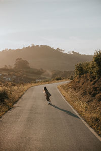 Back view faceless sporty female in trendy wear riding cruiser board along empty asphalt road in summer countryside on sunny day