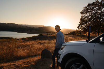 Man standing against car at lakeshore during sunset