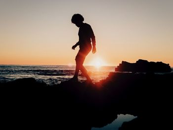 Silhouette person on rock formation by sea against clear sky during sunset