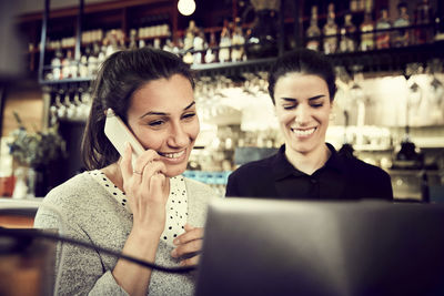 Smiling female coworker talking through smart phone while standing by owner in cafe