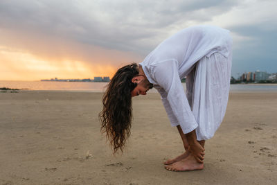 Full length of man exercising while standing at beach against clouds