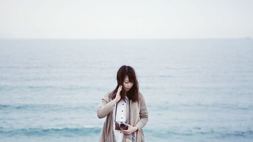 Woman looking down while holding camera against sea at beach