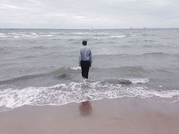 Rear view of man walking in water at beach against cloudy sky