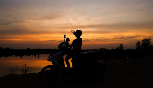 Silhouette boy drinking water while sitting on motor scooter against sky during sunset