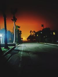 Illuminated street by silhouette buildings against sky at night