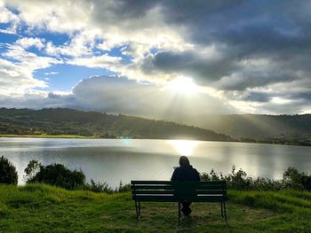 Rear view of man sitting on bench by lake against sky