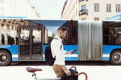 Side view of businessman using smart phone while standing with bicycle against bus on street