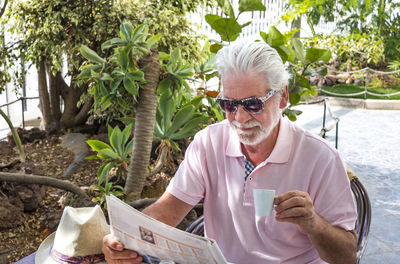 Senior man wearing sunglasses reading newspaper while sitting at cafe