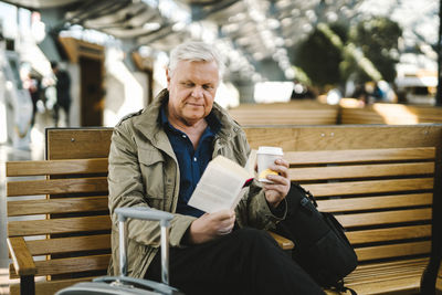 Senior businessman holding disposable cup reading book while sitting on bench at railroad station food court