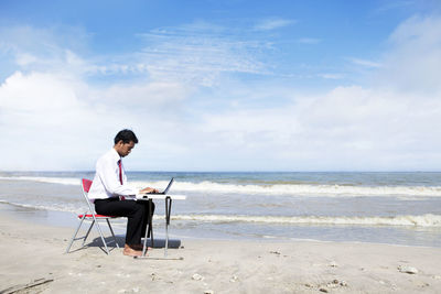 Businessman using laptop while sitting at beach against sky