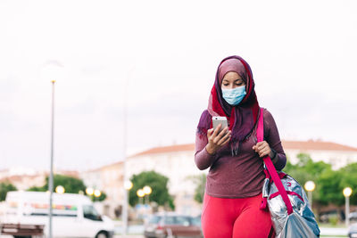 Happy arab sportswoman in hijab walking on street and chatting on mobile phone after training