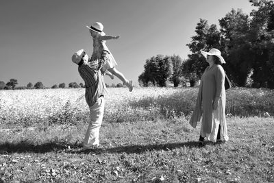 Woman looking at man holding aloft daughter on field against clear sky