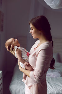 Mother in a white robe holds a small newborn daughter against the wall in the bedroom