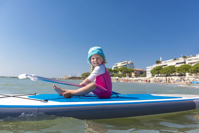 Full length portrait of happy girl paddleboarding on sea against clear blue sky