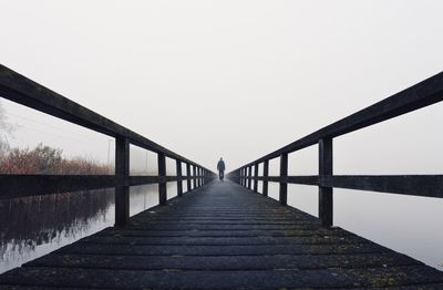 Rear view of man walking on footbridge over lake against sky during foggy weather