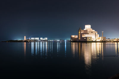 Illuminated buildings by sea against clear sky at night